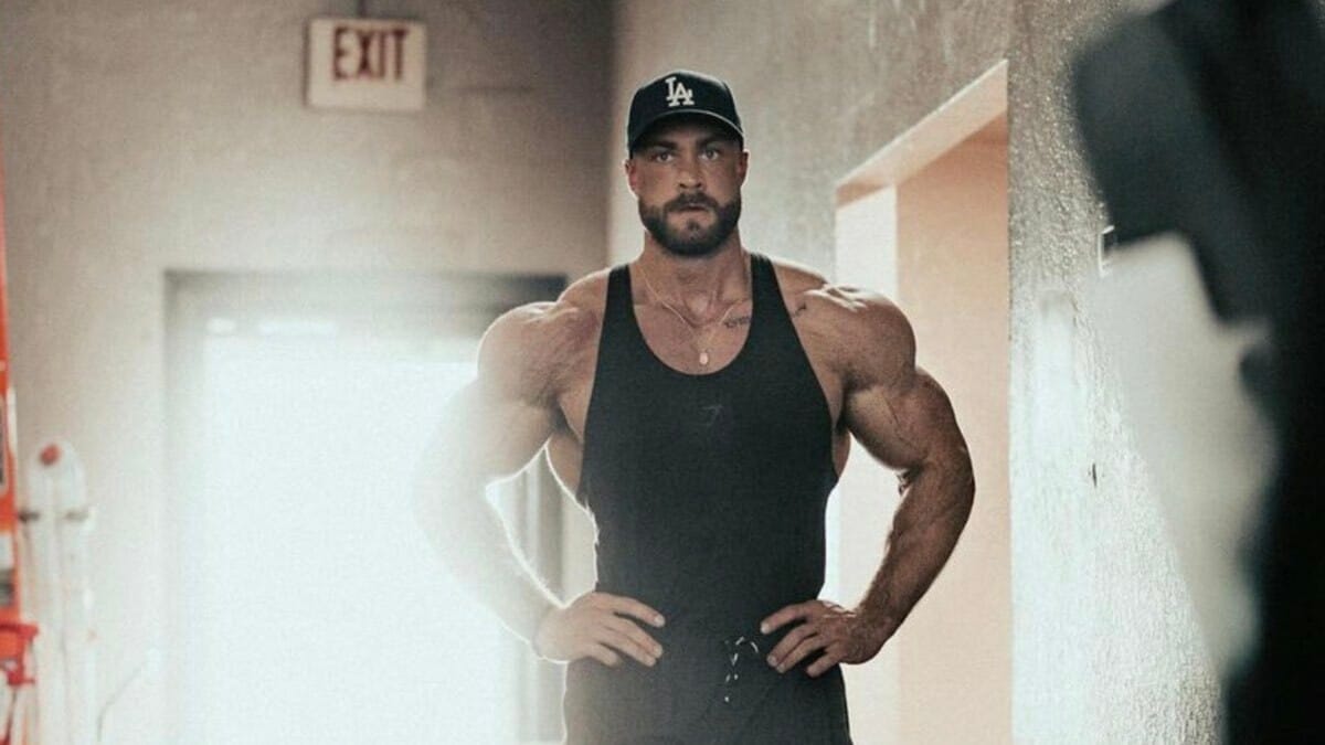 Chris Bumstead with his arms at his side in an open hallway. 